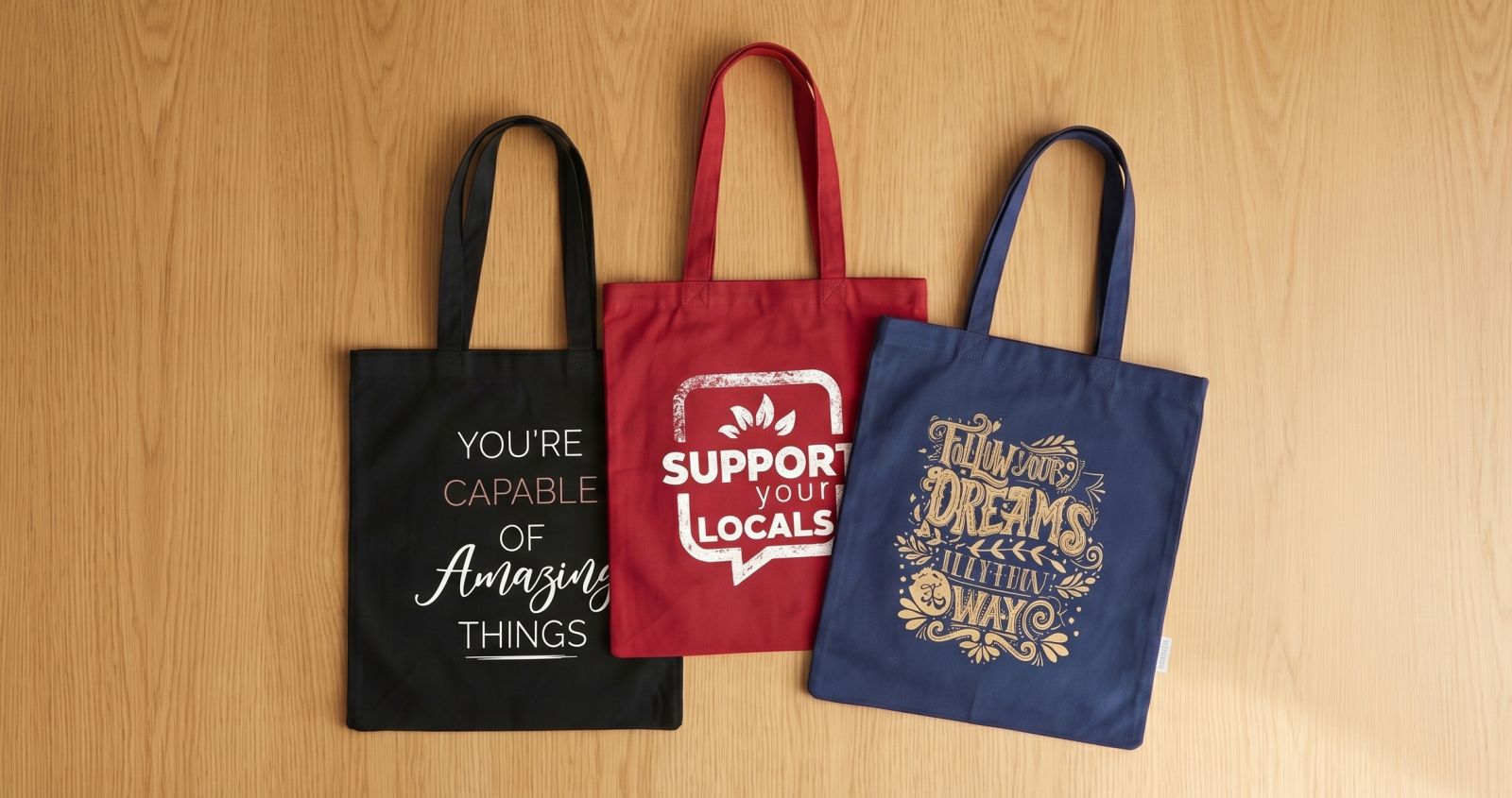 Black, red, and blue tote bags with designs lying on a light brown table.